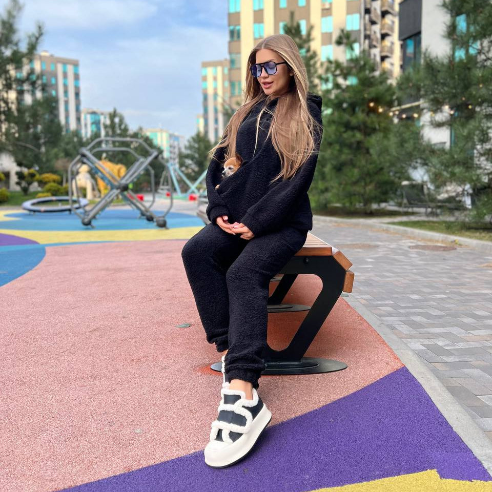 Woman in a beige hoodie pet carrier and pants sitting on a wooden bench outdoors.