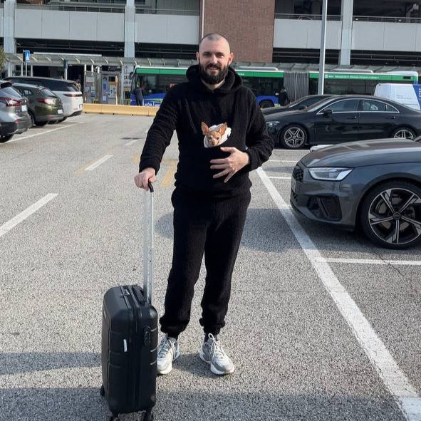 Man in a pet carrier hoodie standing in a parking lot with a building in the background