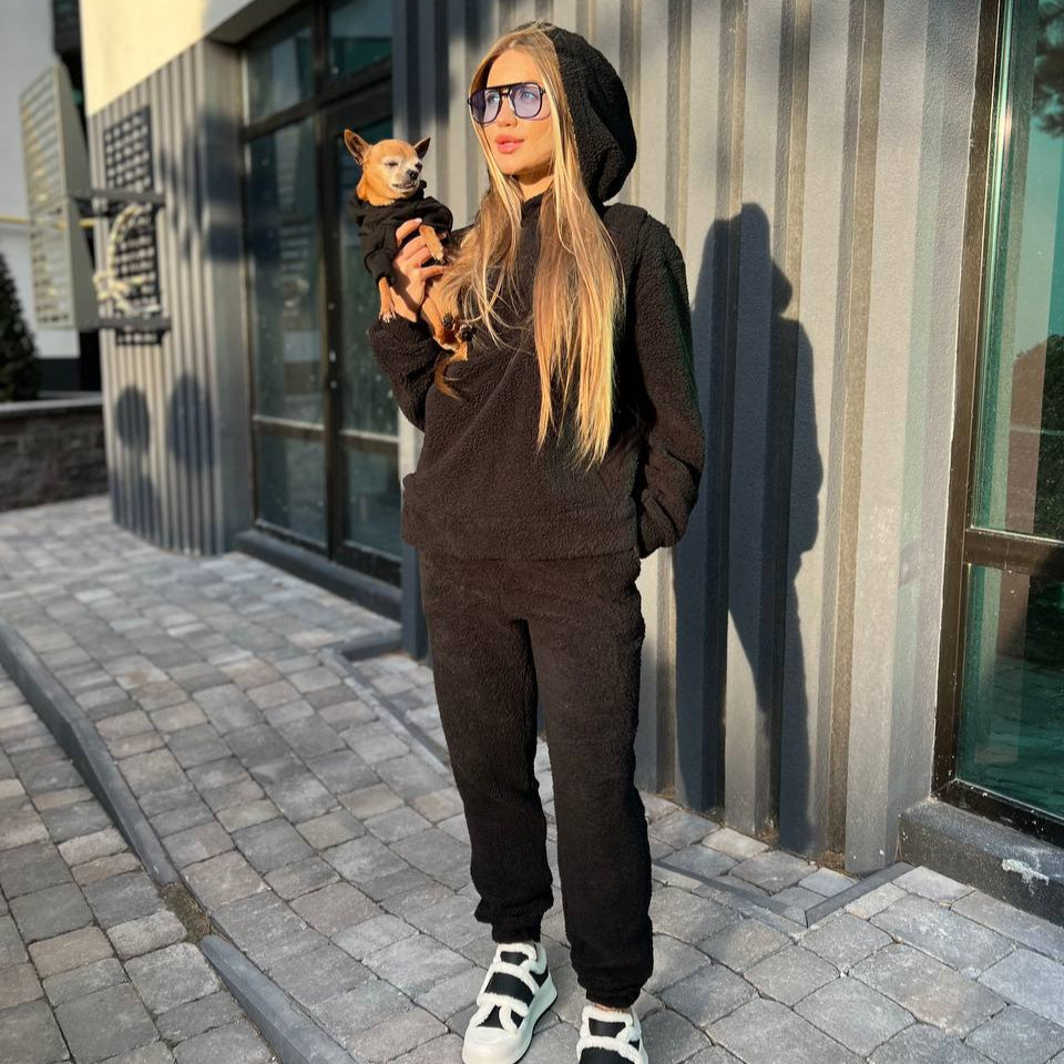 Person wearing a black hoodie pet carrier and pants holding a small dog, standing on a sidewalk.